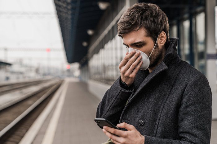 man wearing face mask at transit station, using smart phone - immunocompromised
