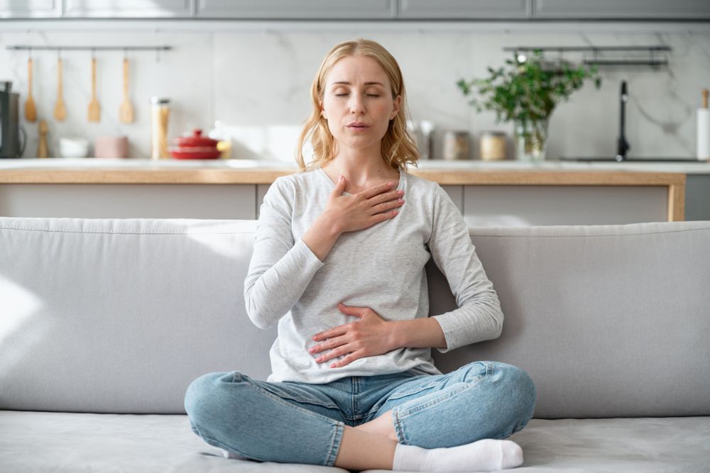 Woman sitting cross-legged on a couch, practicing deep breathing with one hand on her chest and one on her abdomen, appearing calm and focused.