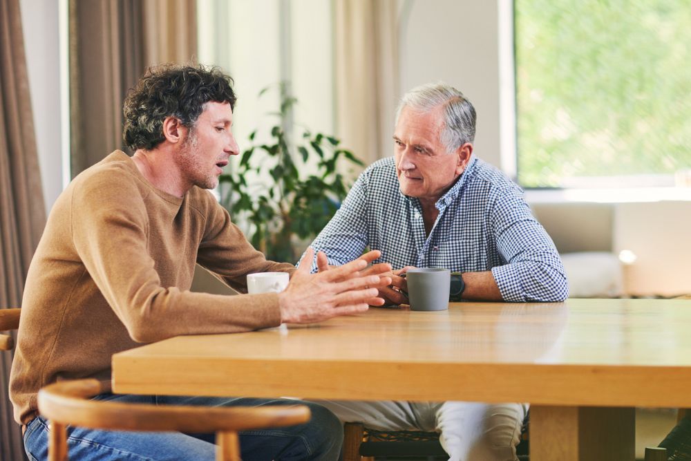 Two men sitting at a table having a serious conversation over coffee, with one man speaking and gesturing while the other listens attentively.