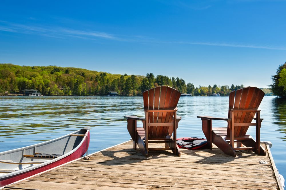Two wooden chairs and a canoe on a dock overlooking a calm lake, surrounded by trees—evoking relaxation, nature, and peaceful summer moments.