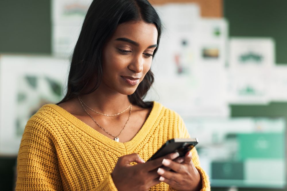 A young woman wearing a yellow sweater smiles while using a smartphone, suggesting engagement with a wellness or recovery app.