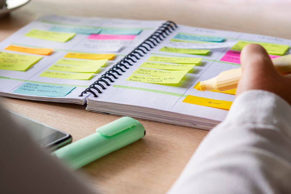 A person highlights a section in an open planner filled with colorful sticky notes and schedules. A green highlighter and smartphone rest on the desk nearby.