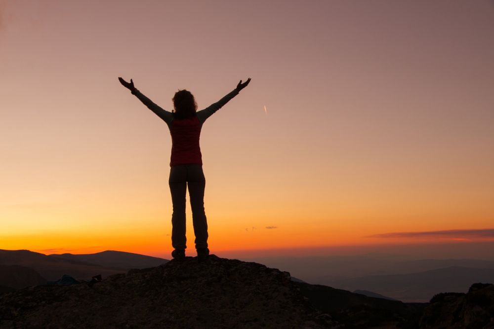Silhouetted person standing on a mountaintop at sunset with arms raised in triumph, symbolizing empowerment, achievement, and personal transformation.
