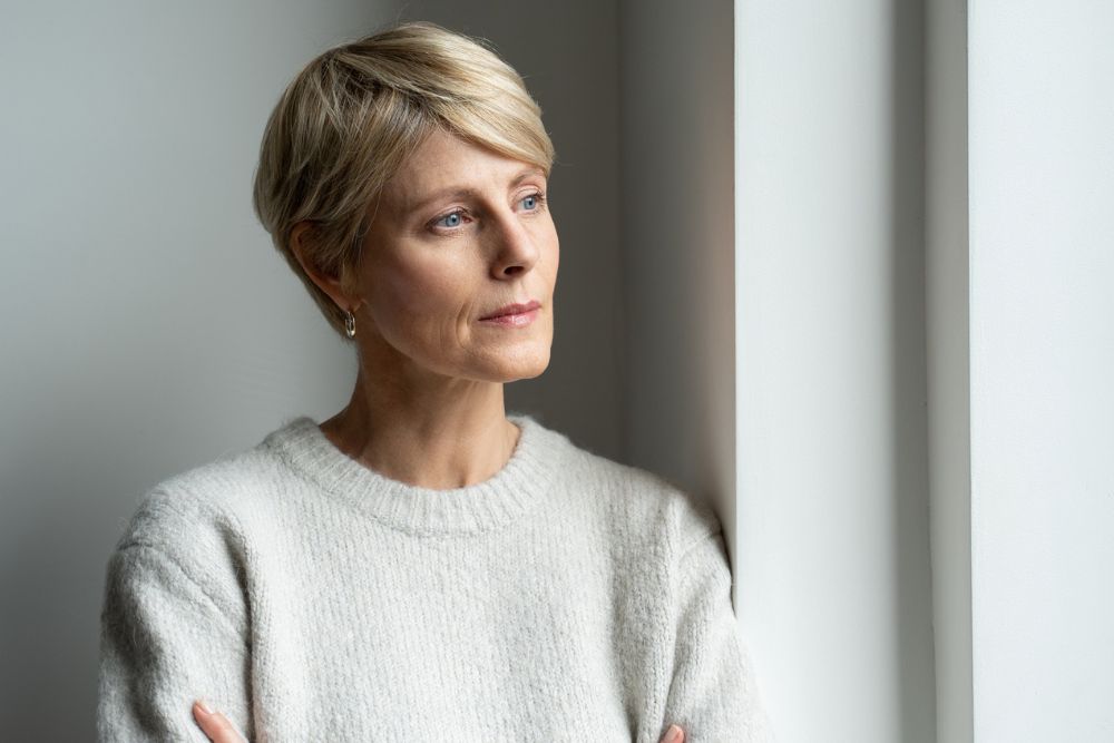 A thoughtful woman with short blonde hair, wearing a light sweater, stands by a window and gazes outside with a contemplative expression.