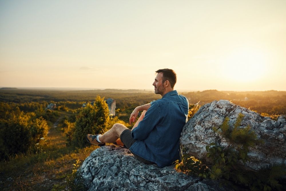 A man sits on a rocky hill at sunset, looking out over a vast landscape of trees and fields, appearing peaceful and reflective.