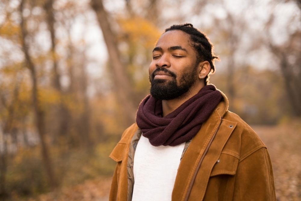 Man standing outdoors with eyes closed and a peaceful expression, practicing mindfulness in a wooded area during autumn.