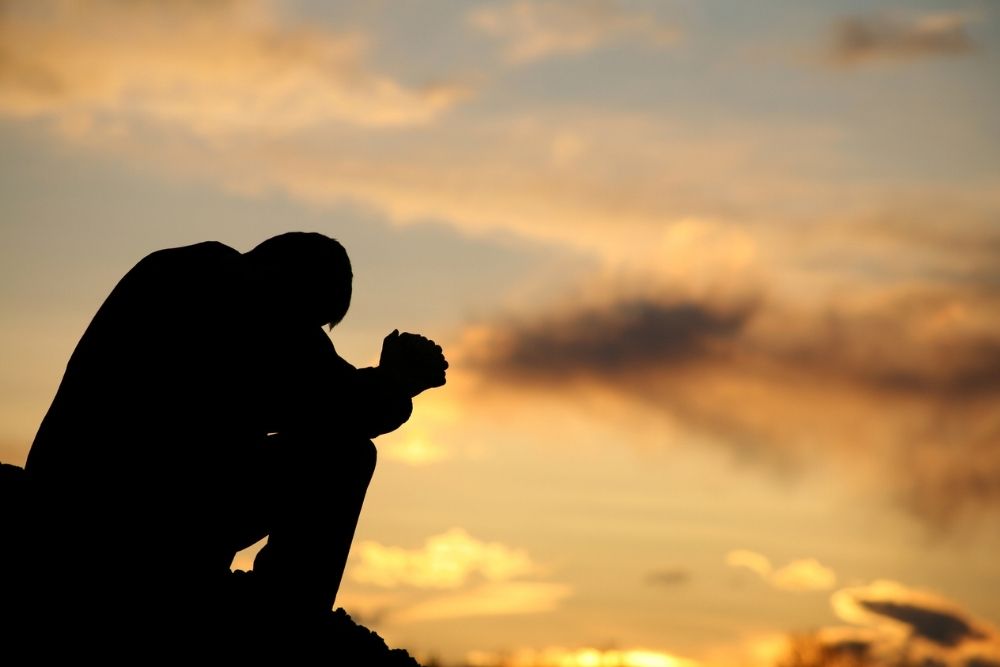 Silhouette of a person sitting with head bowed and hands clasped, appearing reflective or distressed against a dramatic sunset sky.