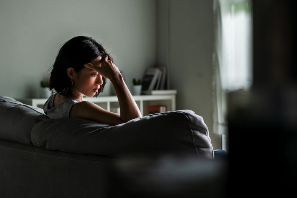 A woman sits on a couch with her head in her hand, looking distressed in a dimly lit living room.