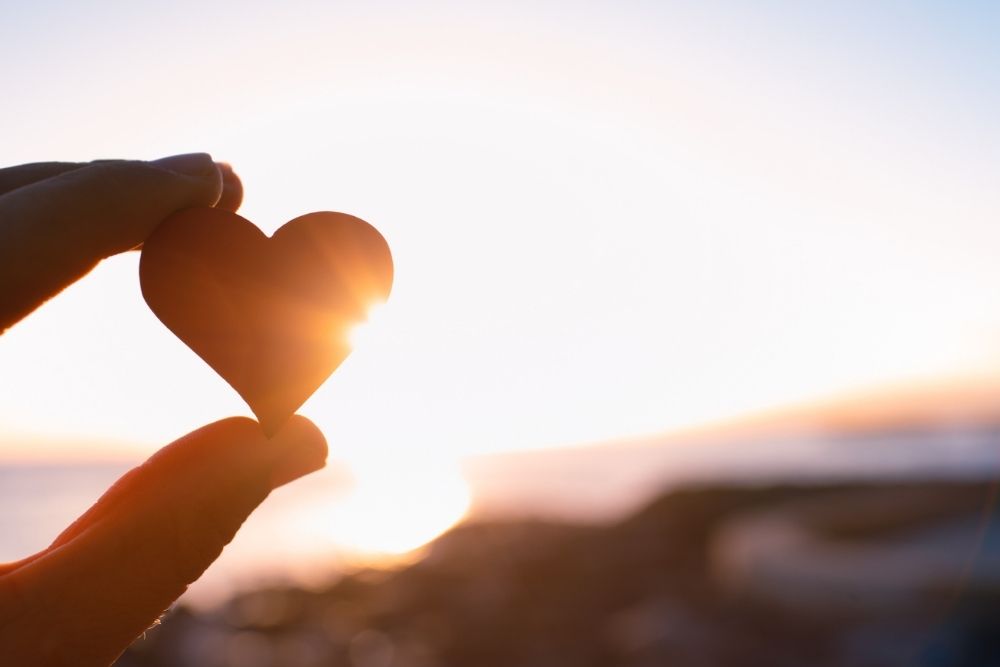 Gratitude A hand holds a small heart-shaped object toward the setting sun, symbolizing love, hope, and gratitude.