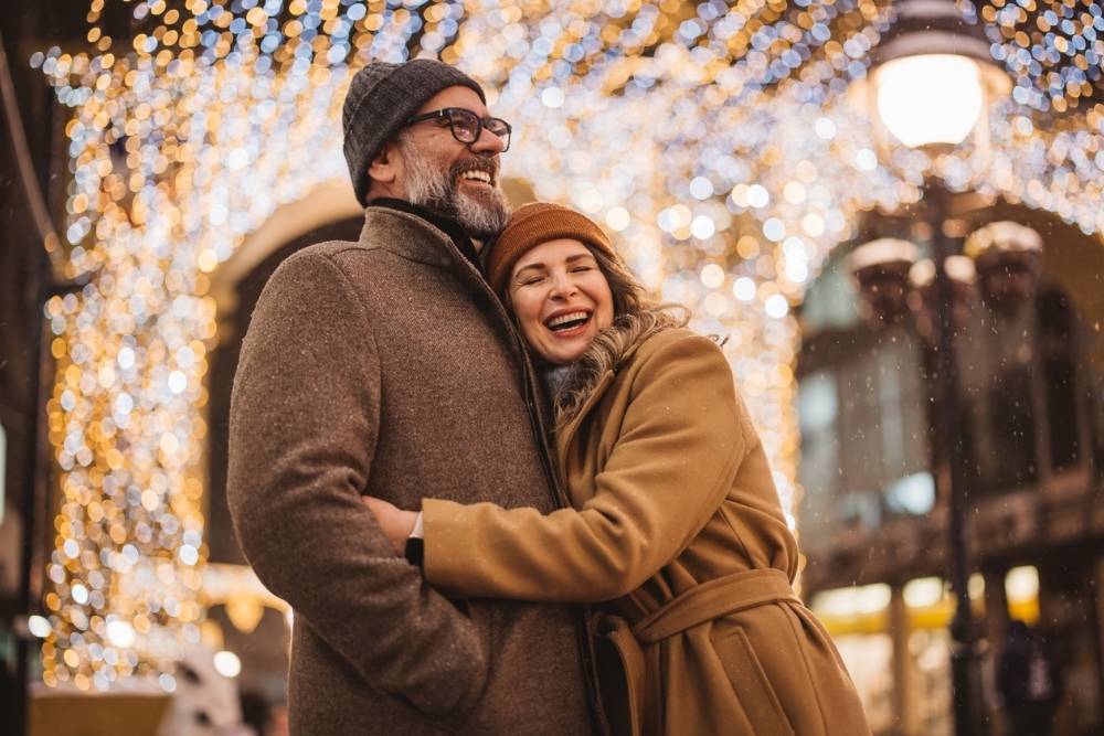 A couple stands outdoors on a winter evening, bundled in warm coats and hats, laughing and embracing under sparkling holiday lights. The festive, glowing background creates a joyful, romantic atmosphere.