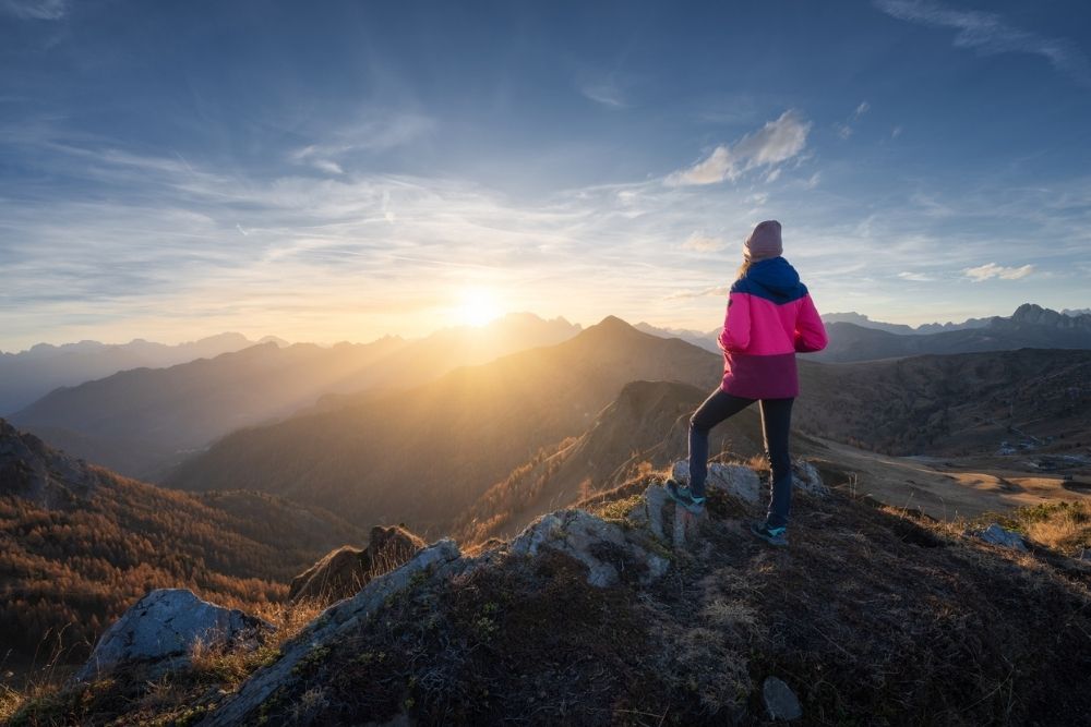 A person standing on a mountain ridge at sunrise, looking out over a vast landscape of mountains and valleys.