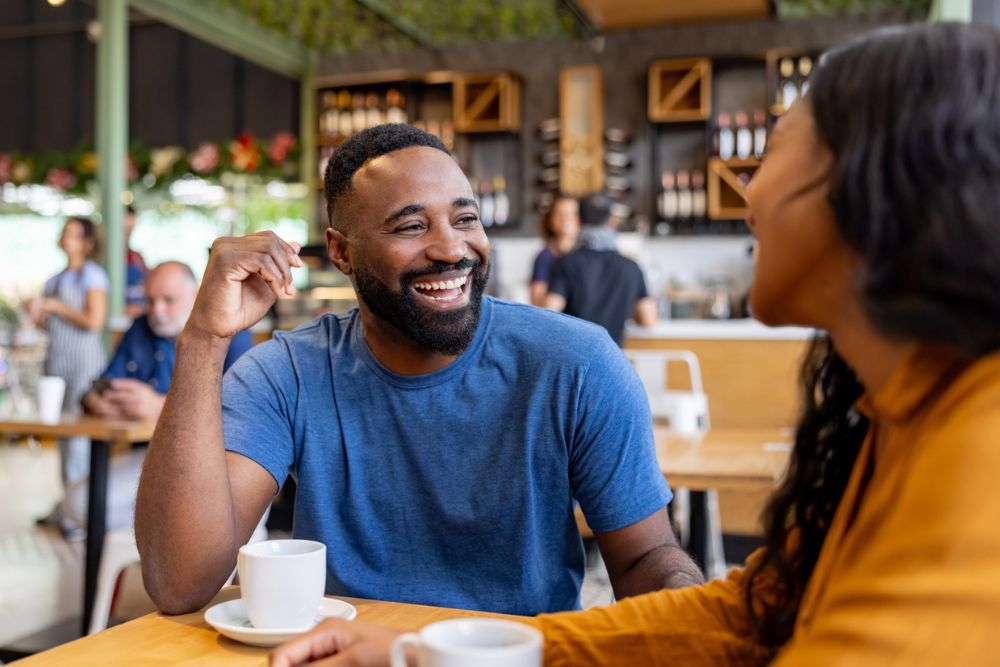 A man smiles and laughs while talking with a friend over coffee in a bright, casual café.