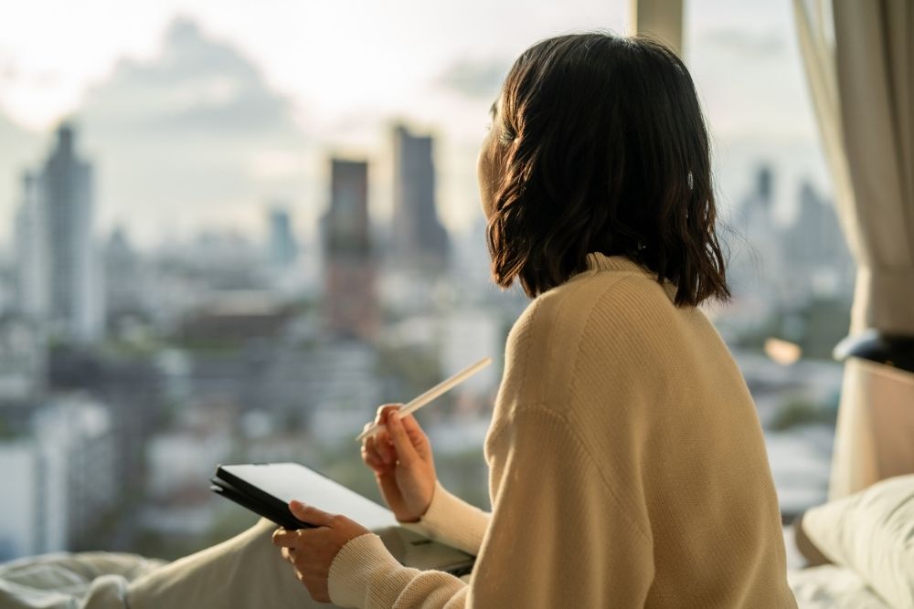 A person sitting by a window, holding a notebook and pen while looking out over a city skyline in a reflective moment.