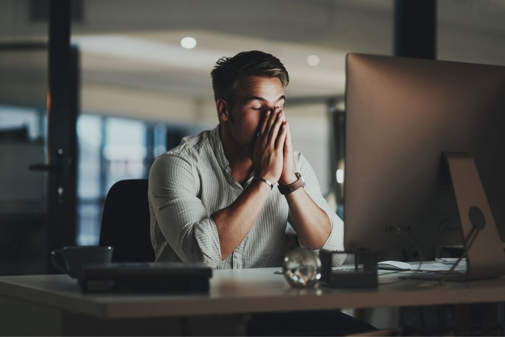 Confidential treatment for professionals is available - A person sits at a desk in a dimly lit office, appearing stressed with hands on face, illuminated by a computer screen, suggesting late-night work.