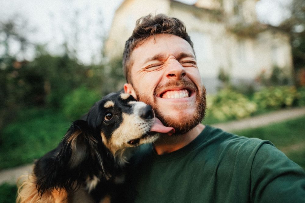 Smiling man takes a selfie outdoors as his dog licks his cheek, capturing a playful moment of joy.
